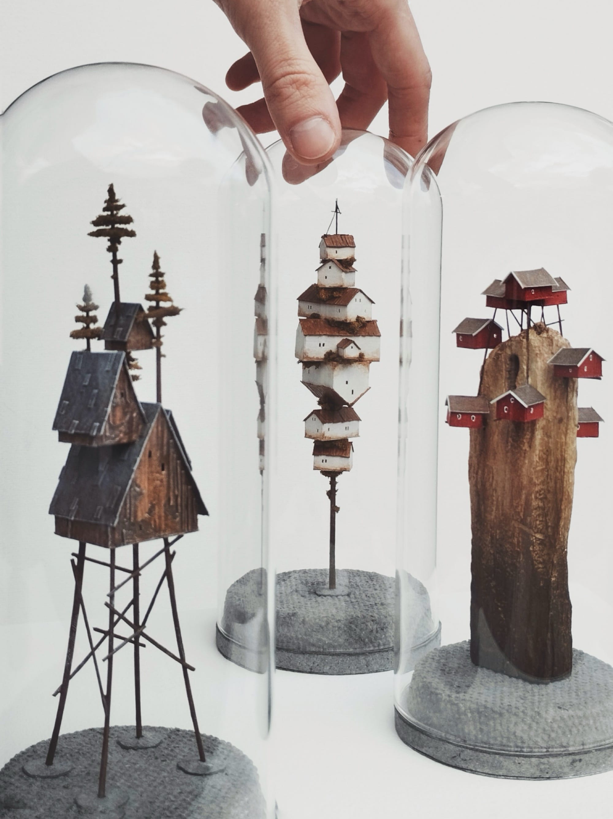 a hand touches one of three glass domes around miniature sculptures of tiny, stacked houses by Michael Davydov