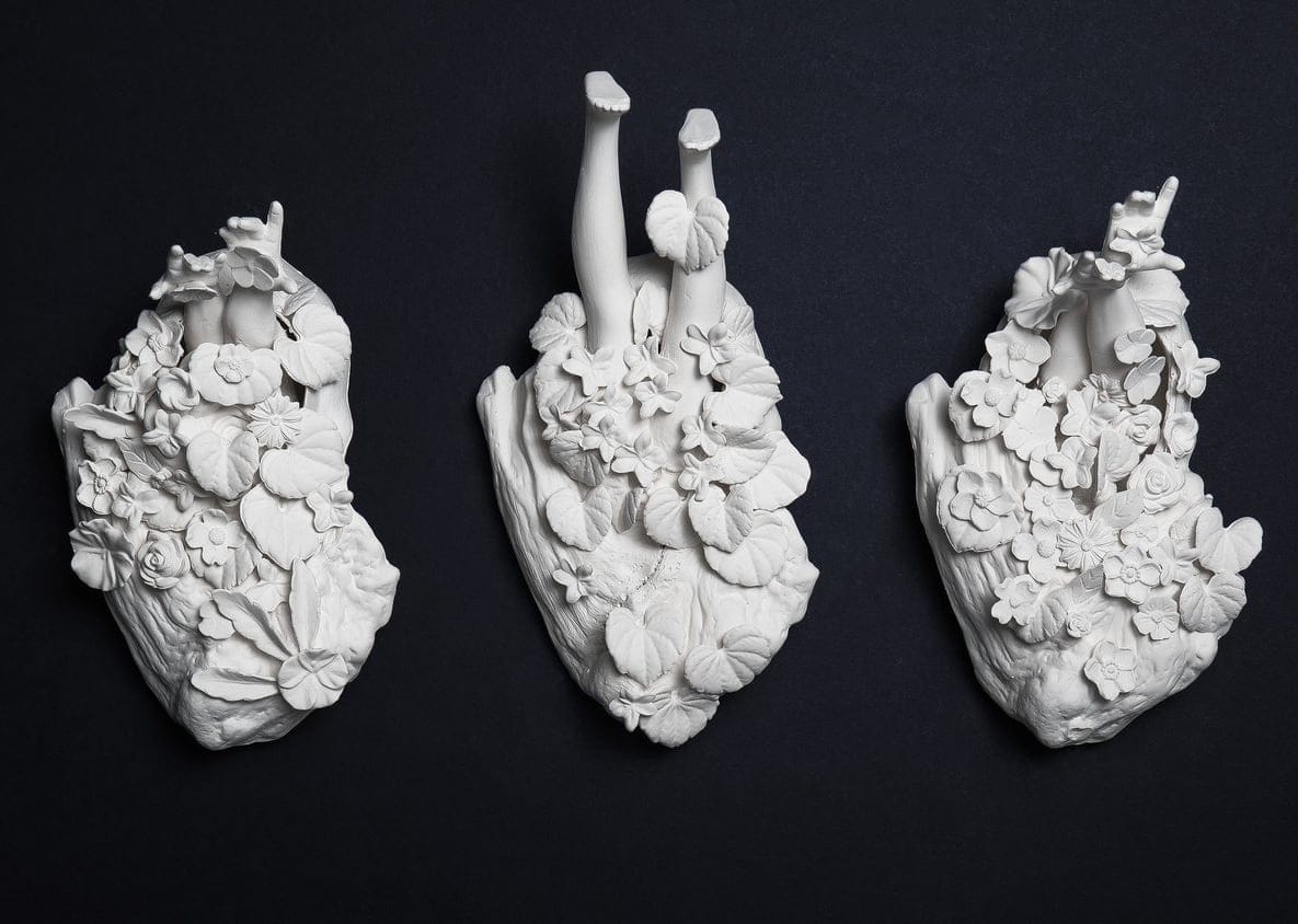 Three white porcelain botanical clusters hang on a wall, each with human limbs jutting from the top