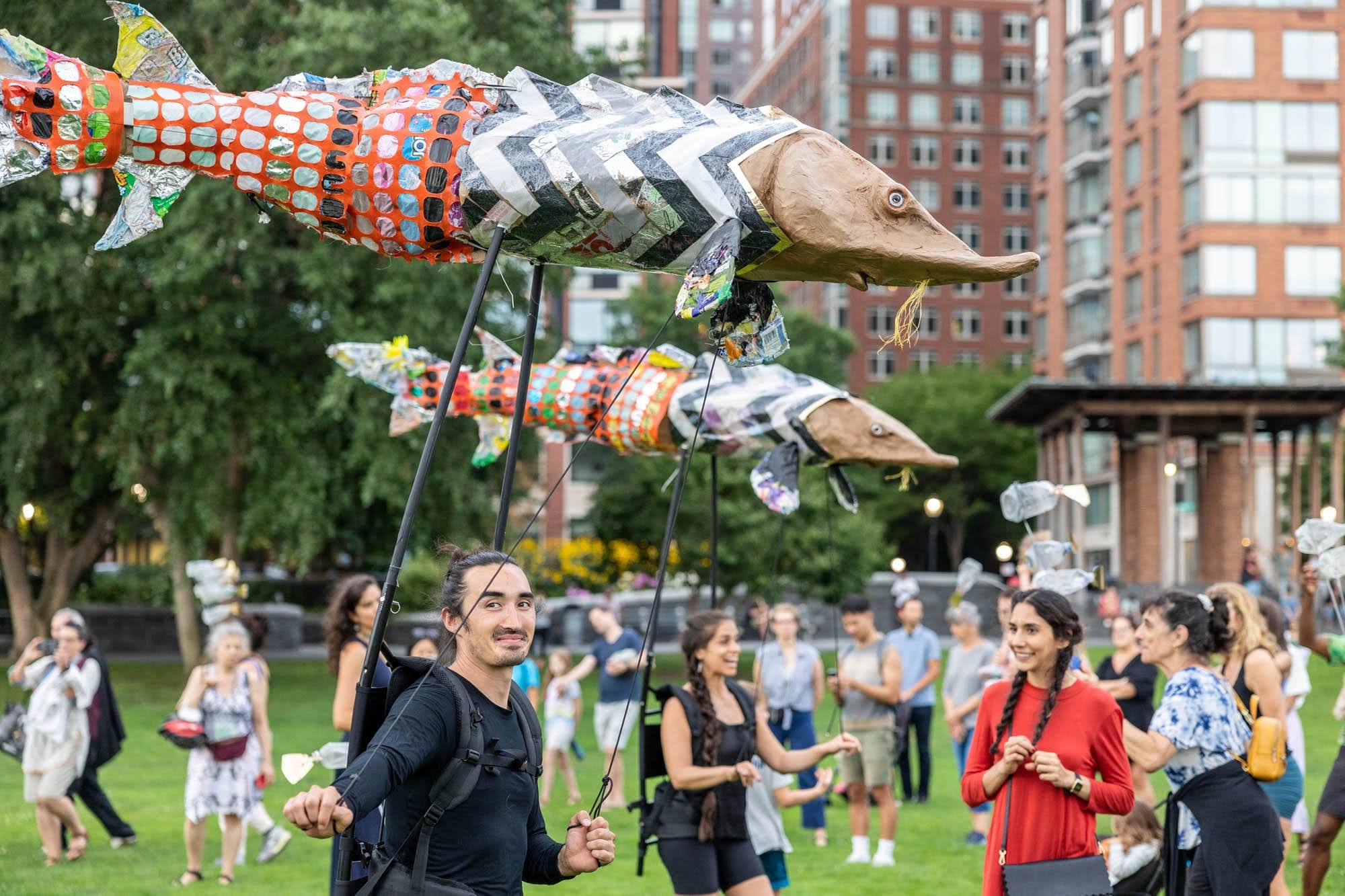 a procession of people in a park carry three puppets of sturgeons on poles