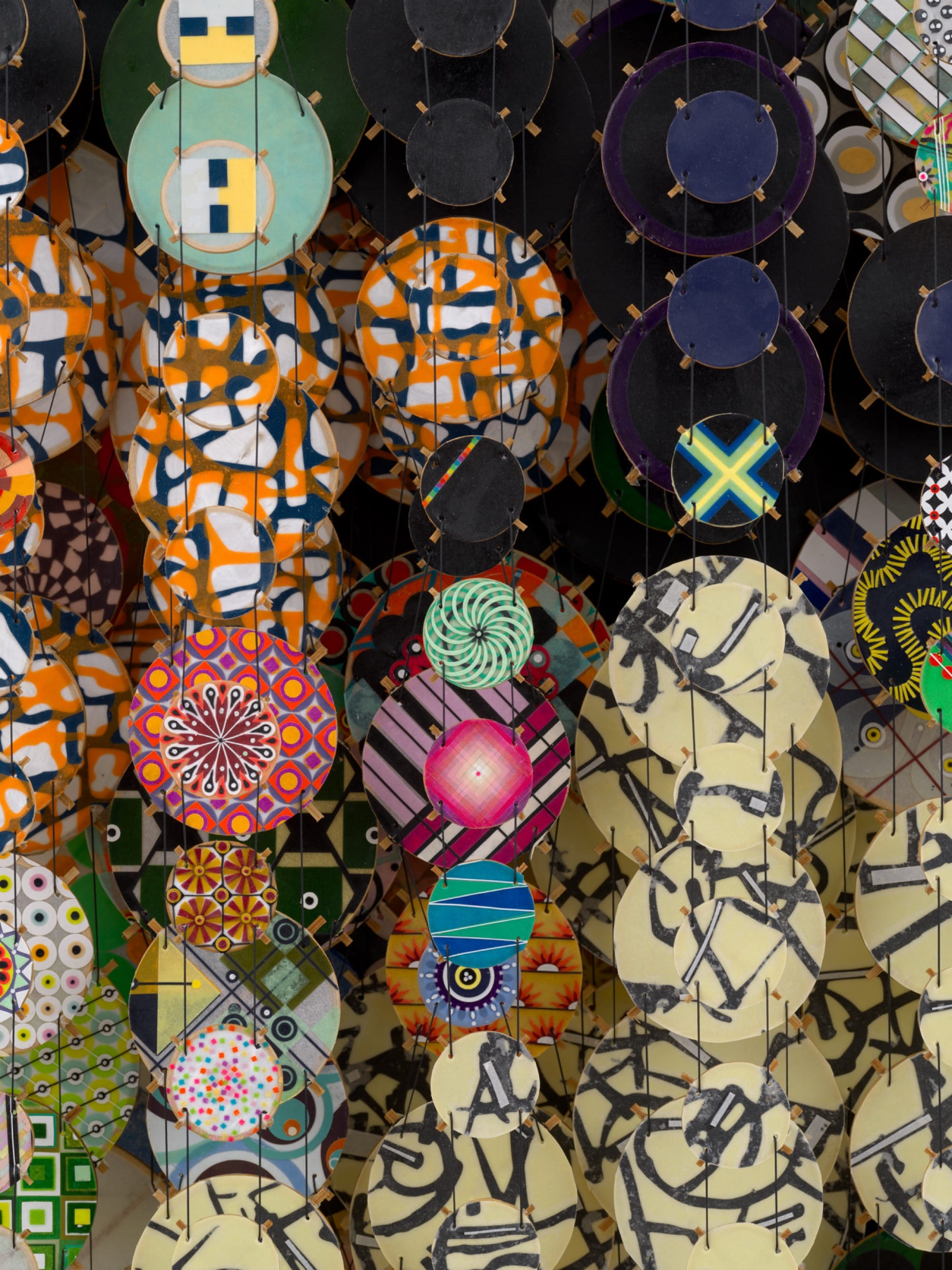 a detail of an abstract artwork by Jacob Hashimoto made from twine and numerous colorful, patterned paper discs installed in rows for a layered effect