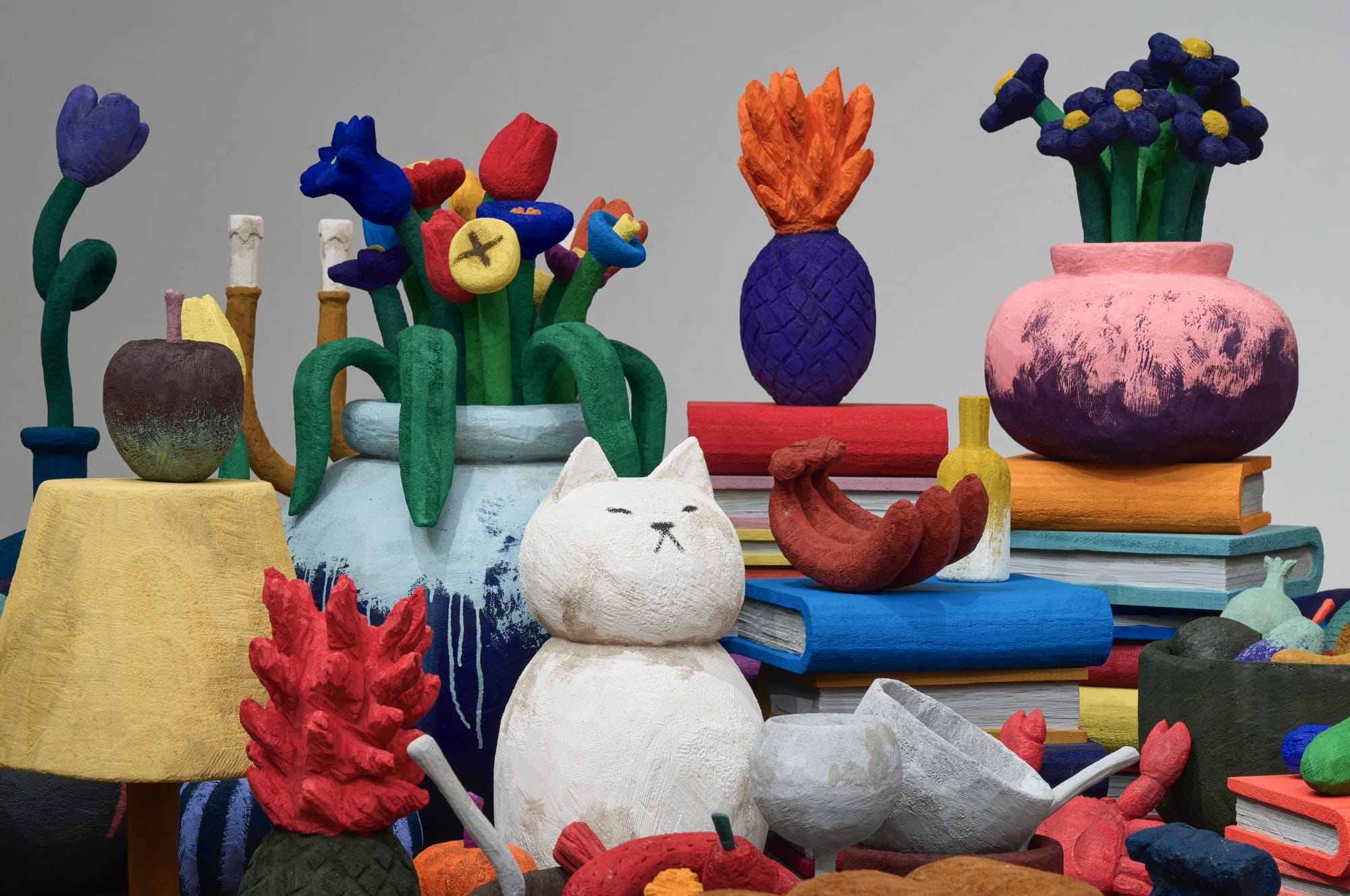 a detail of a sculptural installation by Yuichi Hirako of an oversized table covered with cartoonish domestic forms like vases, food, candlesticks, cats, and other objects