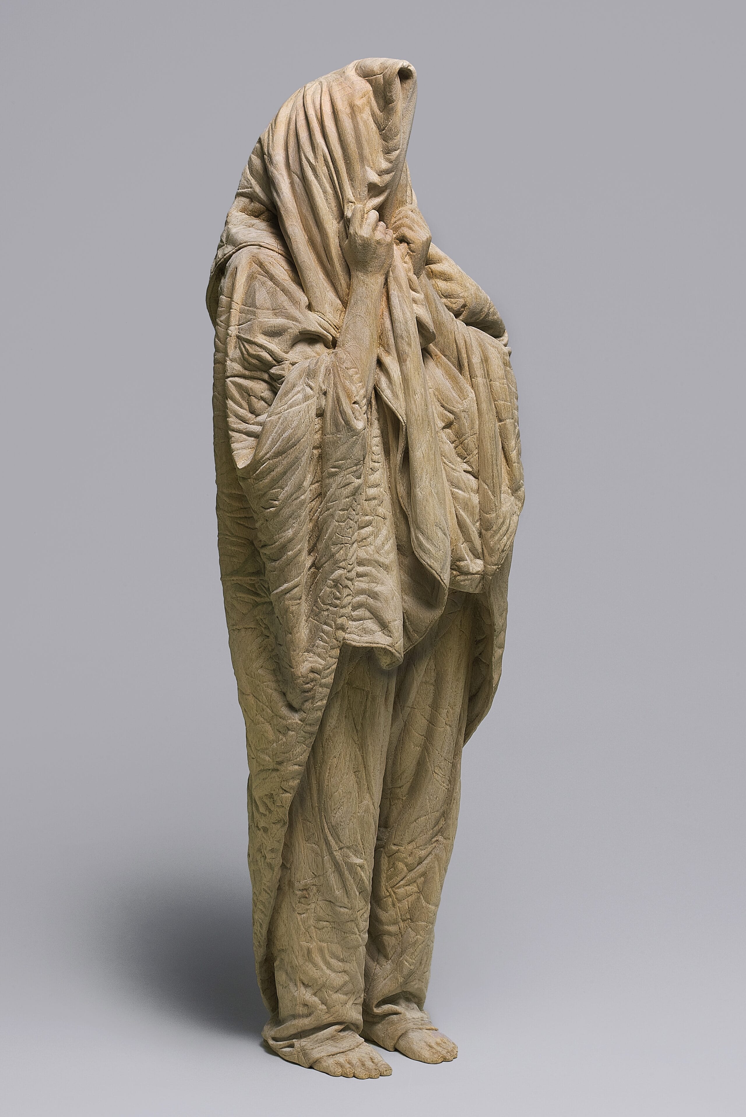 a carved wooden sculpture by Ikuo Inada of a figure standing with a blanket over their head as if very sleepy