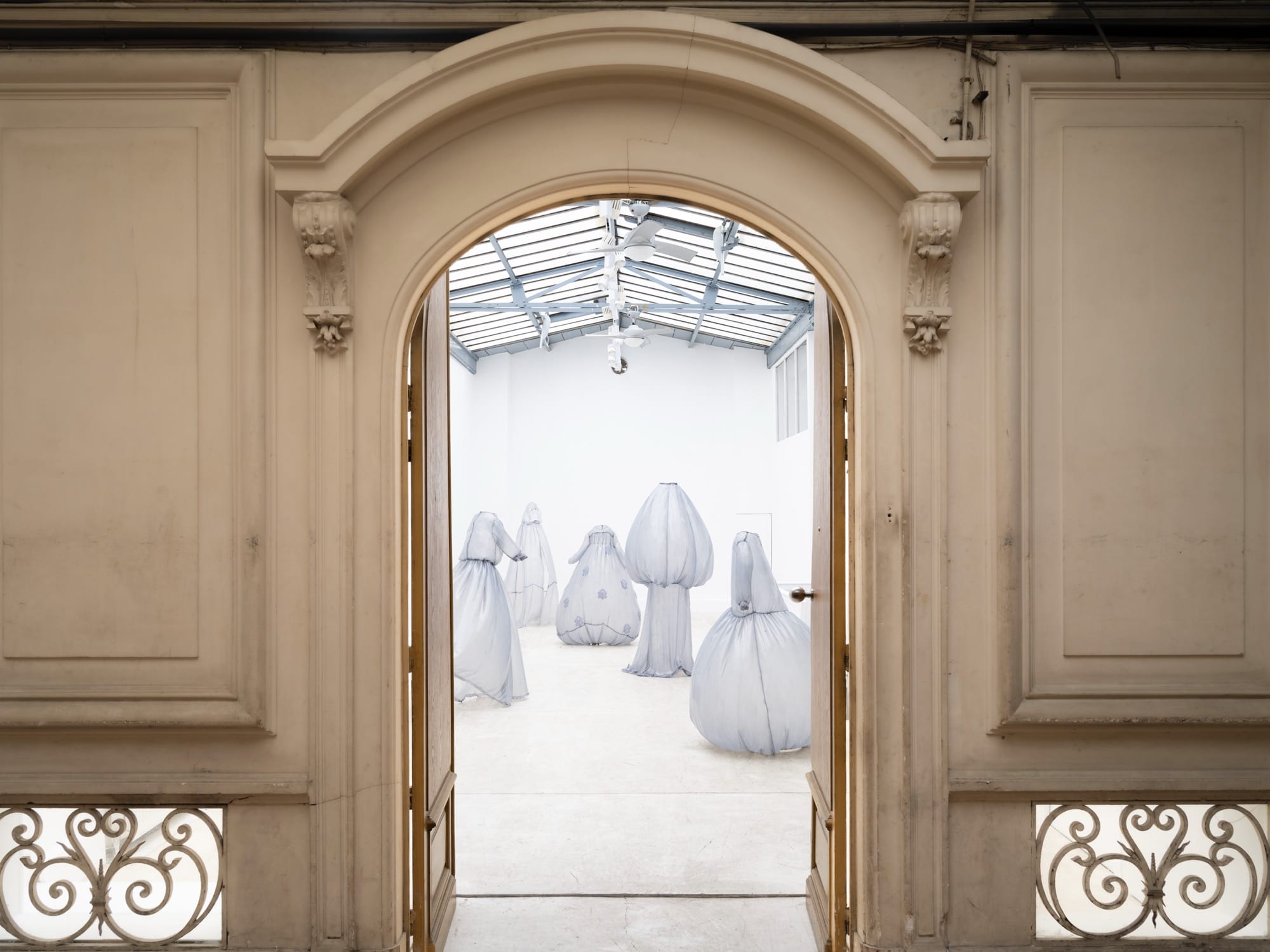 a collection of stiff pale blue fiberglass victorian style dresses by Brandon Morris that stand without wearers in the middle of a gallery, seen through an archway