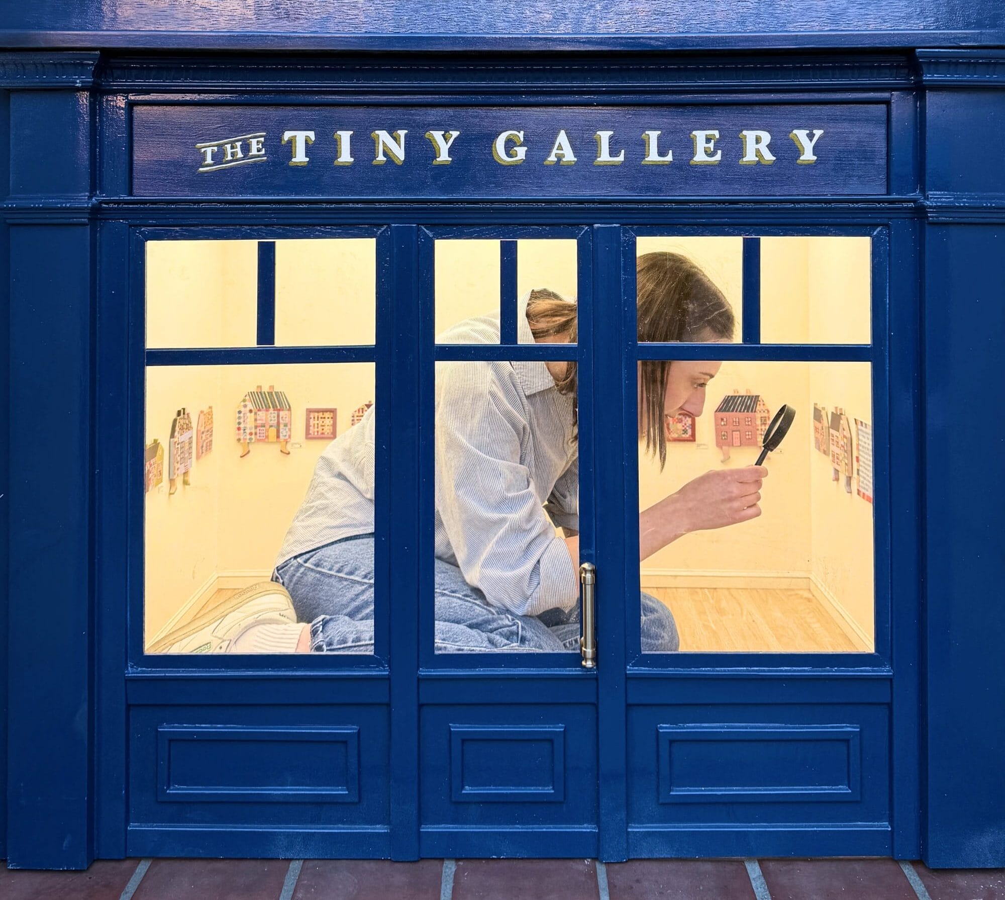 a person kneels inside of a 1/6-scale gallery, holding a magnifying glass up to a painting